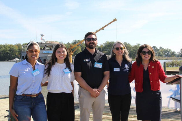 Five people stand side by side outdoors on a dock near water, smiling at the camera with a boat and trees in the background.