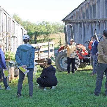 People stand around farm buildings listening to a man in a cap.