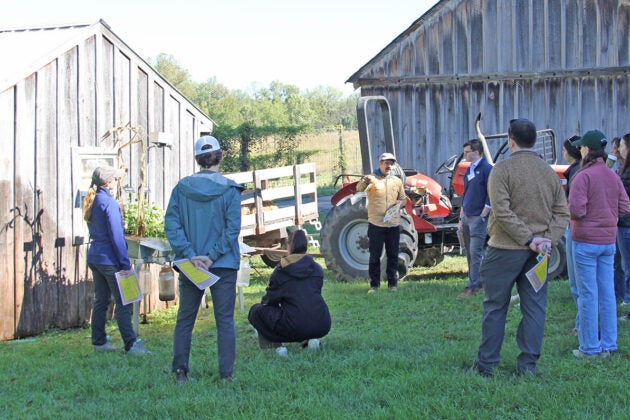 People stand around farm buildings listening to a man in a cap.