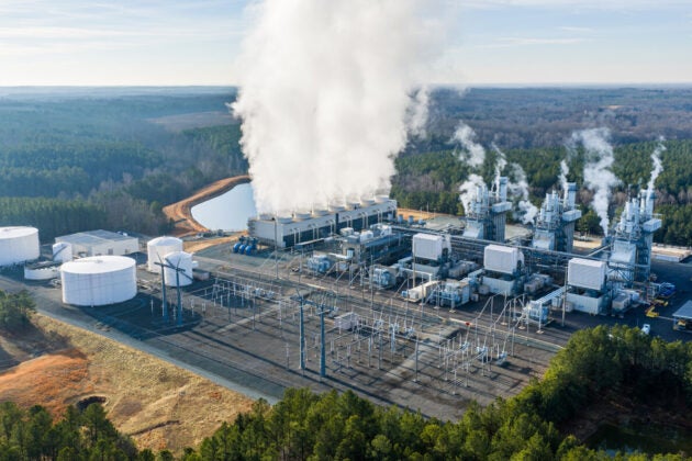 A power plant spewing vapor into the air, set against a background of forests.
