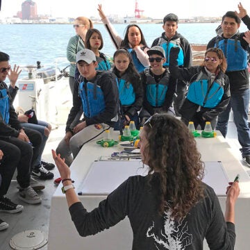 Students circle the edge of a boat listening to a CBF educator at center.