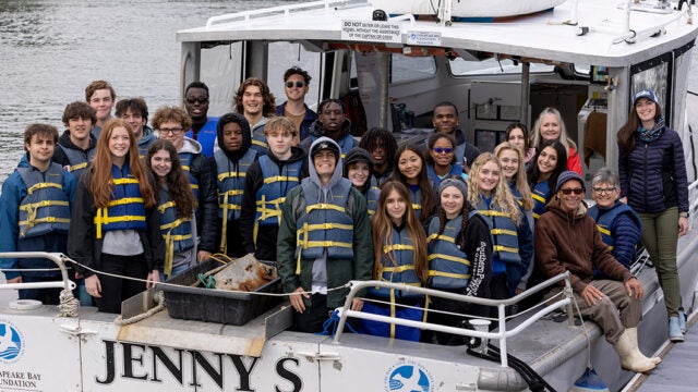 A couple dozen teachers and students stand and smile on the stern of a boat.