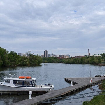A white boat sits at a dock along a river. The Richmond, VA skyline can be seen in the background.