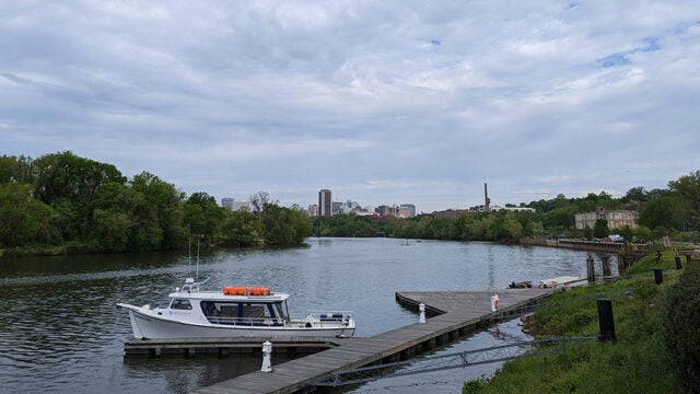 A white boat sits at a dock along a river. The Richmond, VA skyline can be seen in the background.
