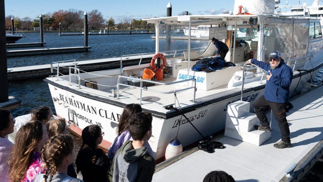 Students stand on a dock while a CBF educator stands on the dock next to a boat talking to them.