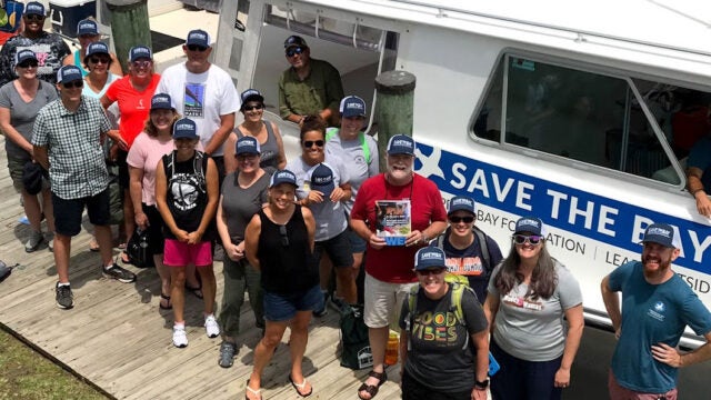 School leaders stand on a dock next to a CBF boat. They smile at the camera while wearing CBF hats.