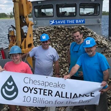 Five volunteers wearing blue hard hats hold a Chesapeake Oyster Alliance banner on a barge loaded with oyster shells