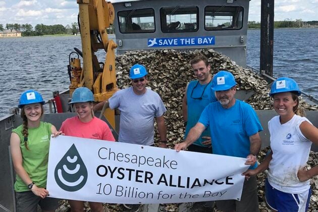 Five volunteers wearing blue hard hats hold a Chesapeake Oyster Alliance banner on a barge loaded with oyster shells