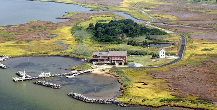 Aerial view of an education center building on a peninsula surrounded by colorful coastal marshes, with a dock, boats, and wetlands featuring bright yellow-green grasses and tidal waterways.