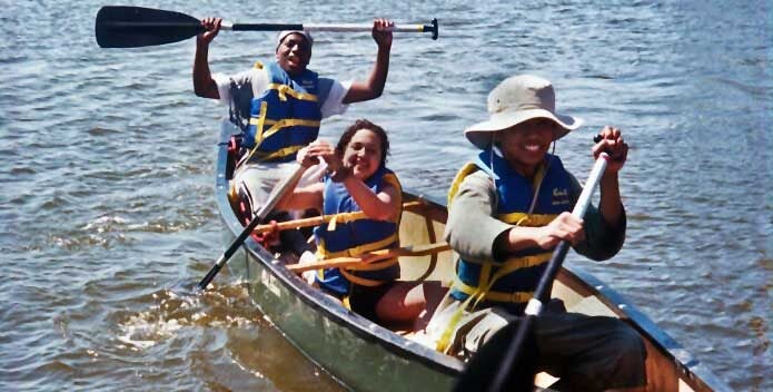 Three students wearing life jackets smile broadly at the camera while paddling a canoe