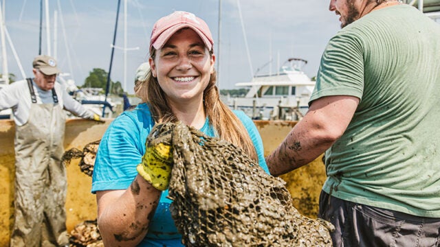 A woman holds a muddy mesh bag of oysters.