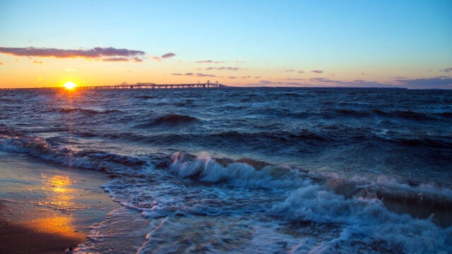 Sunset over a choppy bay with waves crashing onto a sandy shore, and a long bridge stretching across the horizon under a clear blue and orange sky.