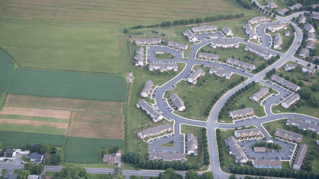 Aerial view of a suburban neighborhood with curved streets and rows of townhouses adjacent to large green agricultural fields. The houses are arranged in clusters with driveways and parking areas, while the farmland shows different shades of green and brown.