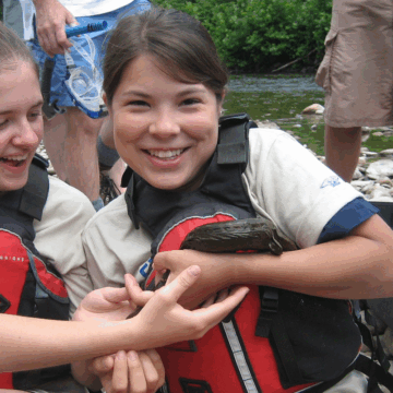 Two people wearing life jackets sit on a rocky riverbank, gently holding an Eastern hellbender. Other individuals stand nearby, and equipment cases are visible in the background near the water.