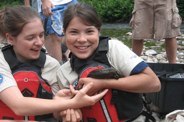Two people wearing life jackets sit on a rocky riverbank, gently holding an Eastern hellbender. Other individuals stand nearby, and equipment cases are visible in the background near the water.