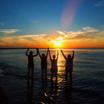 Four people stand in shallow ocean water with arms raised, silhouetted against a vibrant sunset on a scenic beach.