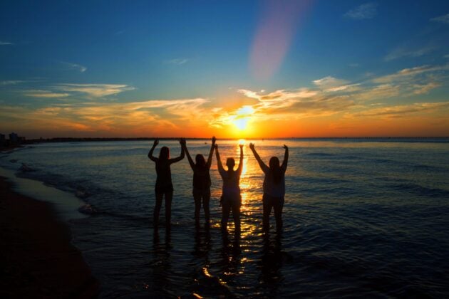 Four people stand in shallow ocean water with arms raised, silhouetted against a vibrant sunset on a scenic beach.