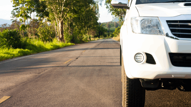 Close-up of the front right side of a white SUV parked on a paved road lined with green trees under bright sunlight.