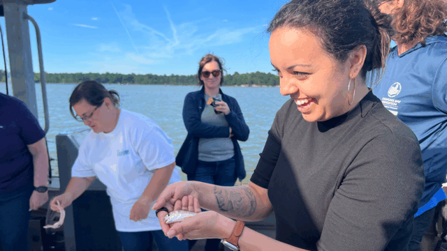 Person holding a small fish on a boat with several others nearby; water and tree-lined shore visible in the background under a clear blue sky.