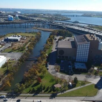 Aerial view of an industrial facility near a waterway, with surrounding roads, bridges, and buildings; a large smokestack and green spaces are visible, and a wide river extends toward the horizon under clear skies.