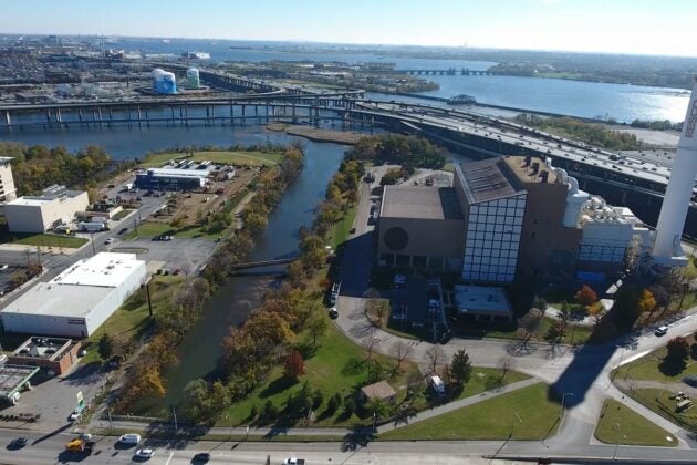 Aerial view of an industrial facility near a waterway, with surrounding roads, bridges, and buildings; a large smokestack and green spaces are visible, and a wide river extends toward the horizon under clear skies.