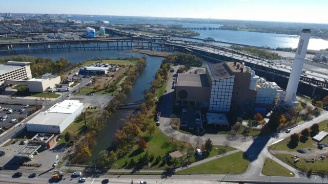 Aerial view of an industrial facility near a waterway, with surrounding roads, bridges, and buildings; a large smokestack and green spaces are visible, and a wide river extends toward the horizon under clear skies.