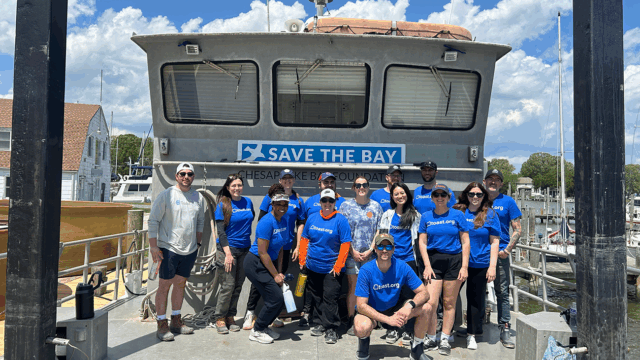 Group of people wearing matching blue shirts standing on a dock in front of a large boat with a sign that reads “Save the Bay” and “Chesapeake Bay Foundation”; marina buildings and boats are visible in the background under a partly cloudy sky.
