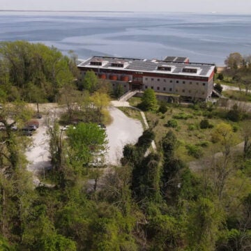 An aerial view shows a building situated between woods and the Bay.
