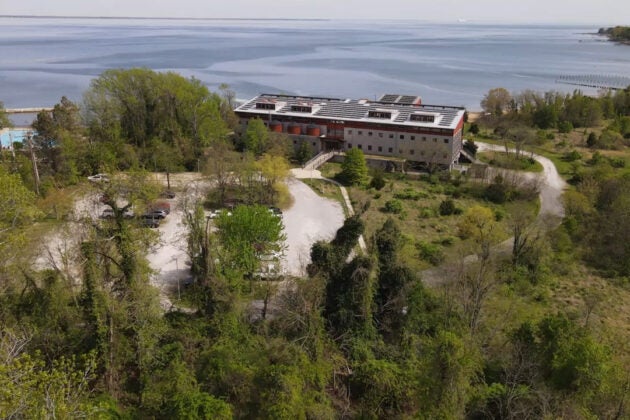 An aerial view shows a building situated between woods and the Bay.