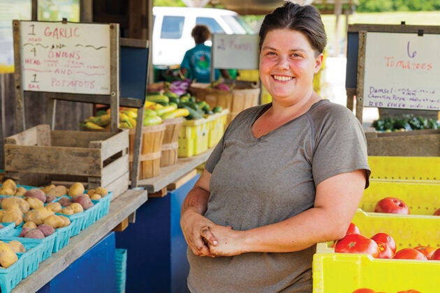 A smiling woman stands in an aisle at a farm stand, surrounded by boxes of tomatoes, potatoes, and garlic.