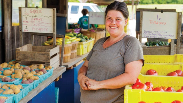 A smiling woman stands in an aisle at a farm stand, surrounded by boxes of tomatoes, potatoes, and garlic.