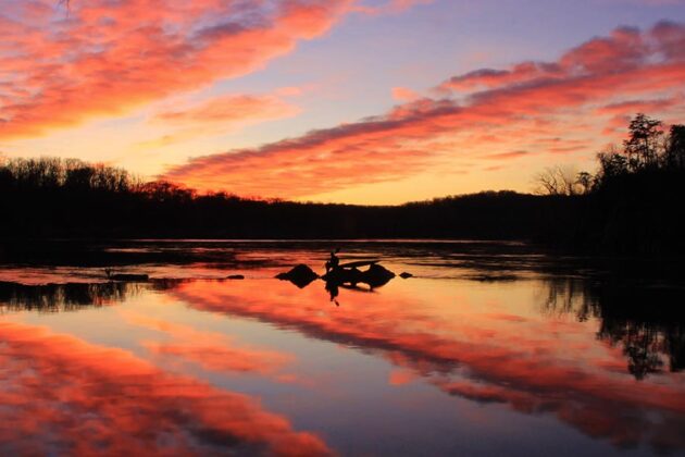An angler fishes from rocks at sunrise with dramatic orange clouds reflecting on calm water, framed by silhouetted trees.