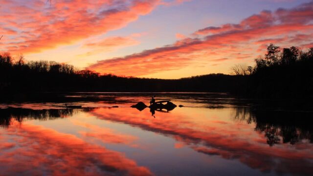 An angler fishes from rocks at sunrise with dramatic orange clouds reflecting on calm water, framed by silhouetted trees.