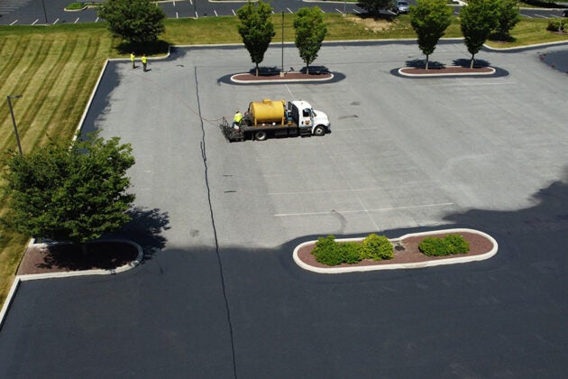 An aerial view of a sealcoating truck with a yellow tank applying pavement sealant in a parking lot with landscaped tree islands.