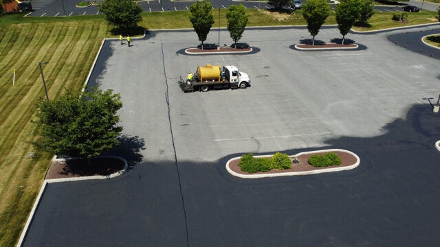 An aerial view of a sealcoating truck with a yellow tank applying pavement sealant in a parking lot with landscaped tree islands.