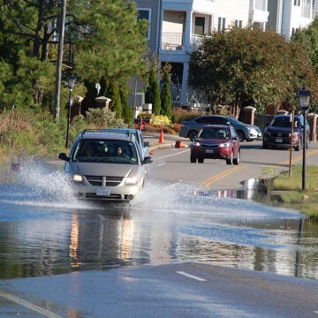 A car drives through floodwater on a street lined with residential buildings and streetlights.