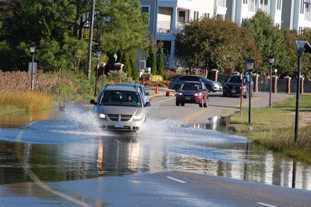 A car drives through floodwater on a street lined with residential buildings and streetlights.