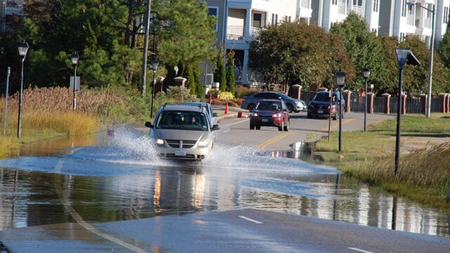 A car drives through floodwater on a street lined with residential buildings and streetlights.