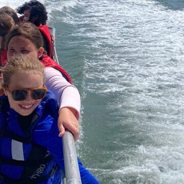 Young students smile as they stand on the side of a boat.