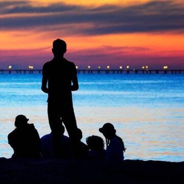 A family is silhouetted in front of the water and an orange sunset.
