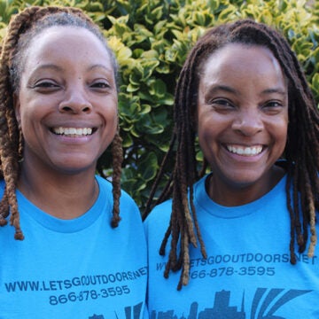 Two Black girls smile next to each other, wearing t-shirts that say www.letsgooutdoors.net.
