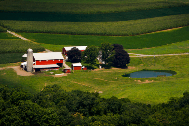 Aerial view of a farm with red barns, a silo, farmhouse, and pond surrounded by rolling agricultural fields.