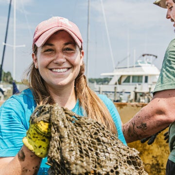 A woman holds a muddy mesh bag of oysters.