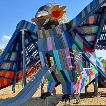 A large rainbow colored bird sculpture has a slide coming out of its body.