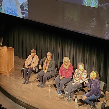 Five women sit on a stage in front of an audience.