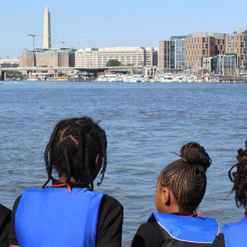Four students in blue life vests look out at the Potomac River with the Washington Monument and DC skyline in the background.