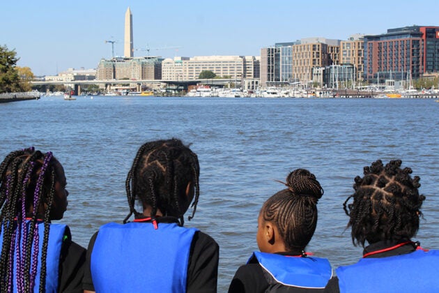 Four students in blue life vests look out at the Potomac River with the Washington Monument and DC skyline in the background.