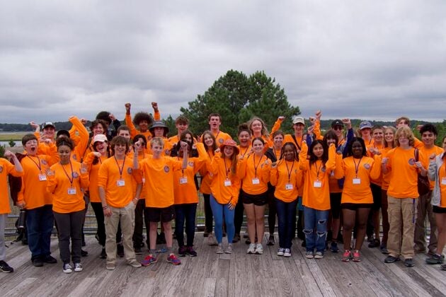 Students in bright orange shirts pose enthusiastically with raised arms on a wooden deck.