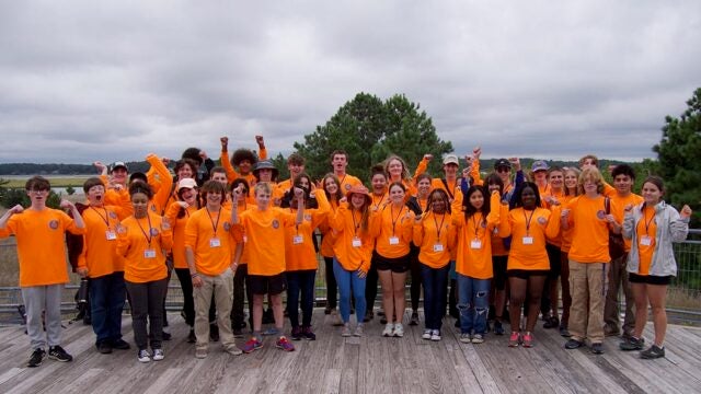 Students in bright orange shirts pose enthusiastically with raised arms on a wooden deck.