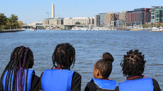 Four students in blue vests look out at the Potomac River with the Washington Monument and DC skyline in the background.